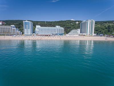 Klares Meer vor breitem Sandstrand mit modernen weißen Hotels und bewaldetem Hügel im Hintergrund.