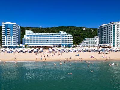 Strand mit goldfarbenem Sand, Sonnenschirmen, Menschen und weißen Hotels vor bewaldetem Hügel.