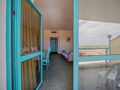 Blick durch offene blaue Tür zu Raum mit Stuhl, Bett und Balkon mit weißen Stühlen und Meerblick.