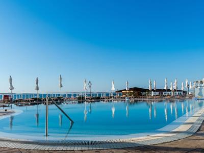 Großer Außenpool mit klarem Wasser, Liegestühlen, Sonnenschirmen und Blick aufs Meer bei blauem Himmel.