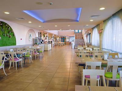 Moderner, heller Restaurantraum mit Tischen, Stühlen, großen Fenstern und blauer Deckenbeleuchtung.