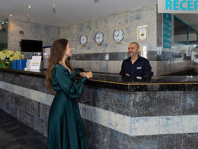 Frau im grünen Kleid spricht an der grauen Hotelrezeption mit einem Mitarbeiter.