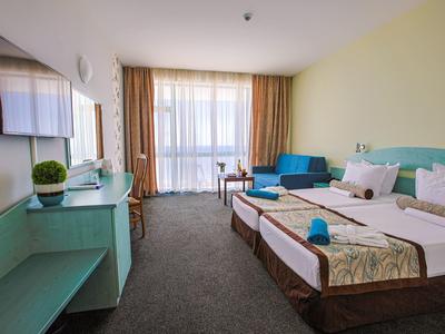 Helles Hotelzimmer mit zwei Einzelbetten, blauem Sofa, großem Fenster und TV an der Wand.
