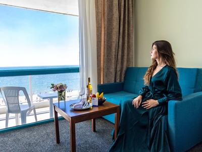 Frau im langen grünen Kleid sitzt auf blauem Sofa und blickt auf Meer durch große Fenster.