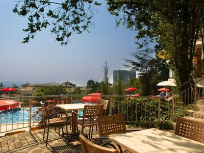 Terrazza esterna con sedie e tavoli che si affaccia su piscina e edifici con ombrelloni rossi.