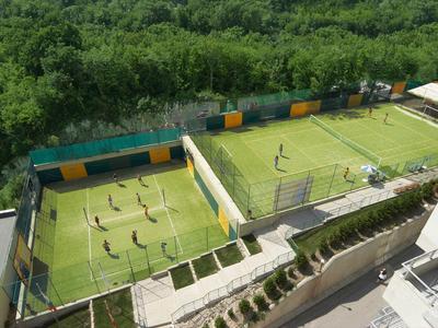 Due piccoli campi da calcio con giocatori, circondati da alberi e edificio adiacente.