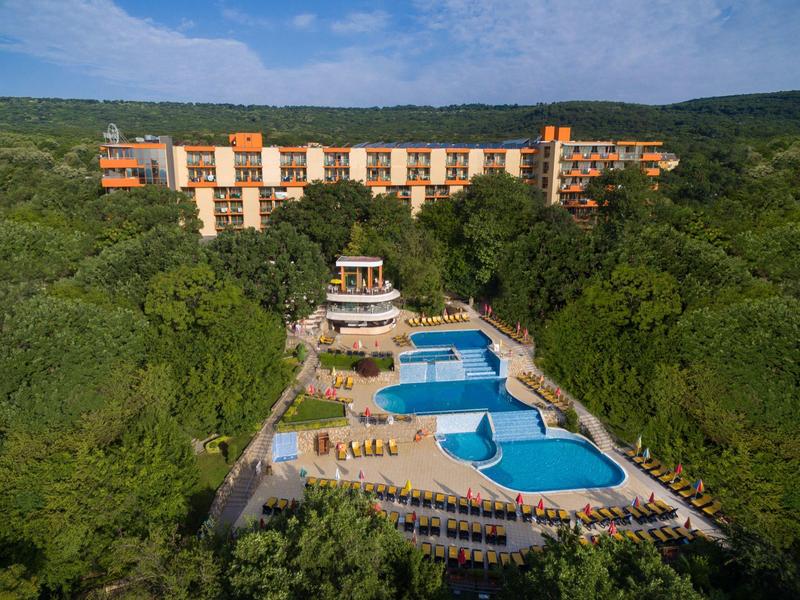 Hotel with large pool and sun loungers, surrounded by dense greenery and wooded hills.