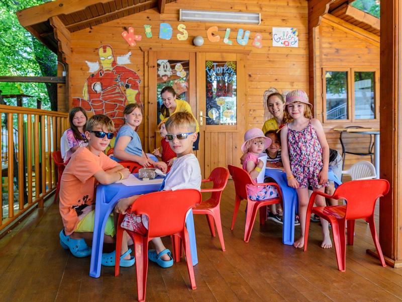 Children sit on colorful chairs on the porch of a wooden house under a 'Kids Club' sign