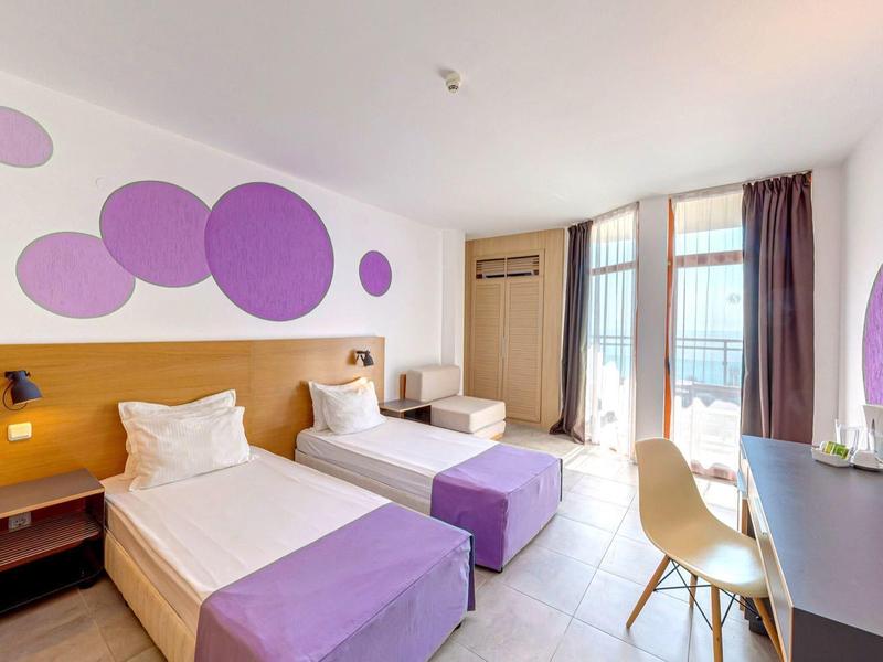 Modern hotel room with two beds, purple accents, and a bright desk by the window.