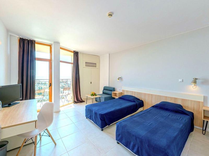 Brightly furnished hotel room with two single beds, desk, and balcony with outdoor view.