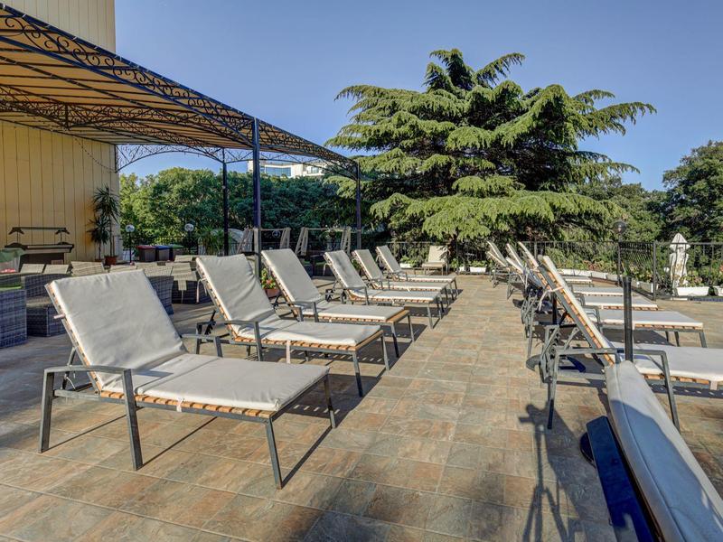 Rząd leżaków na słonecznym tarasie z drzewami w tle.