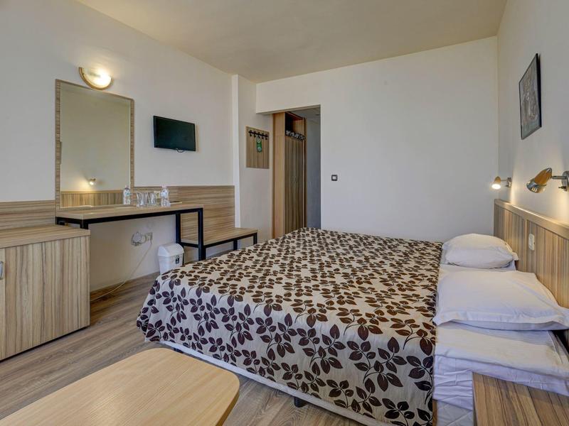 Prosty pokój hotelowy z łóżkiem podwójnym, lustrem ściennym i telewizorem w neutralnych kolorach.