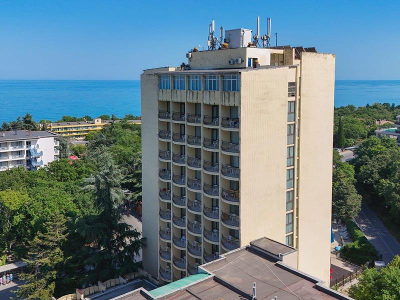 Wysoki budynek hotelu z widokiem na morze i jasne niebo, otoczony drzewami i innymi budynkami.