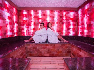 Dwie osoby w białych szlafrokach siedzą w saunie o czerwonym oświetleniu.