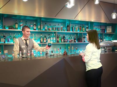 Een vrouw praat met een bartender bij een moderne hotelbar met turquoise verlichte planken.
