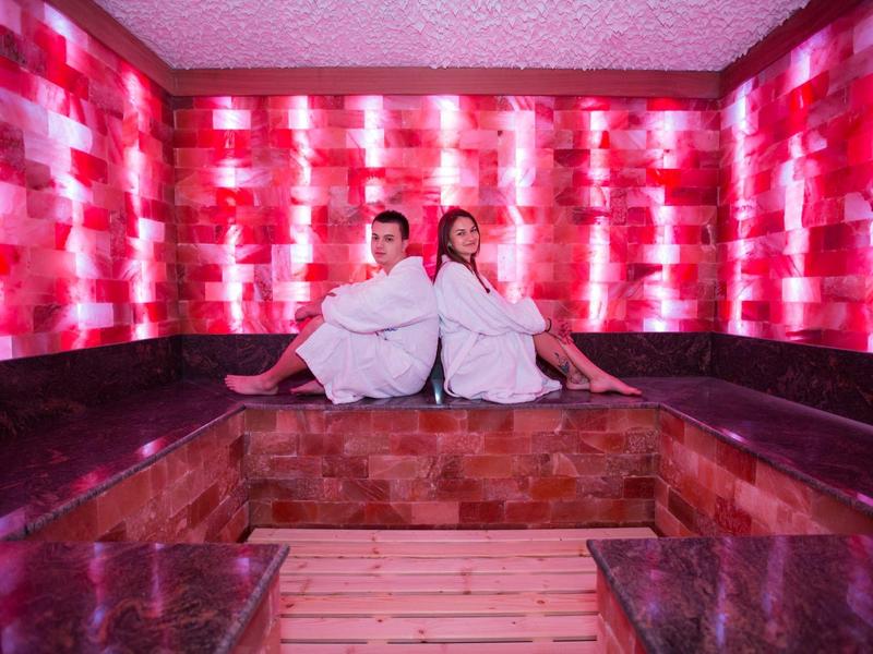 Dwie osoby w białych szlafrokach siedzą w saunie o czerwonym oświetleniu.