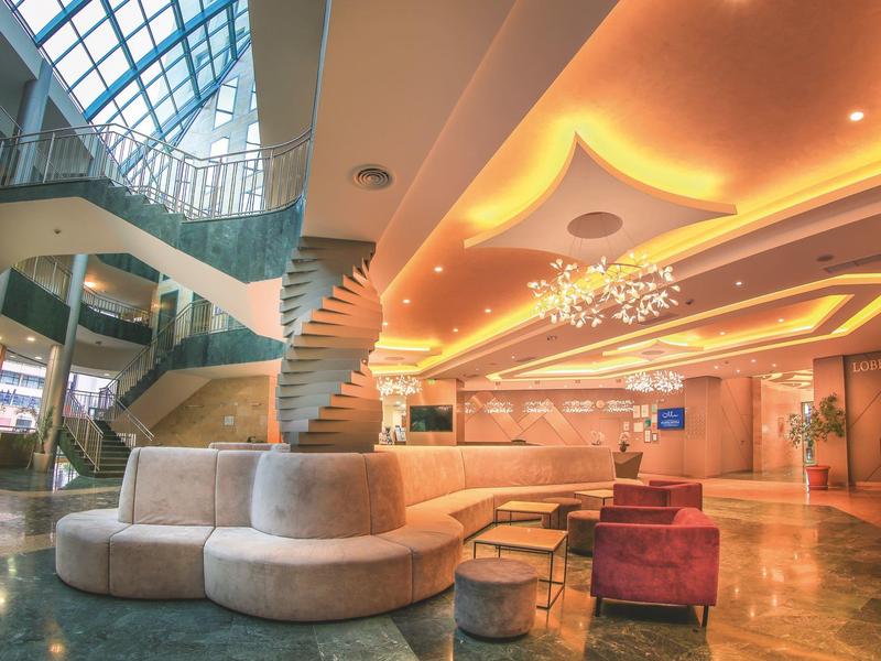 Nowoczesna lobby hotelowa z okrągłą ławką, spiralną kolumną i dużymi oknami.