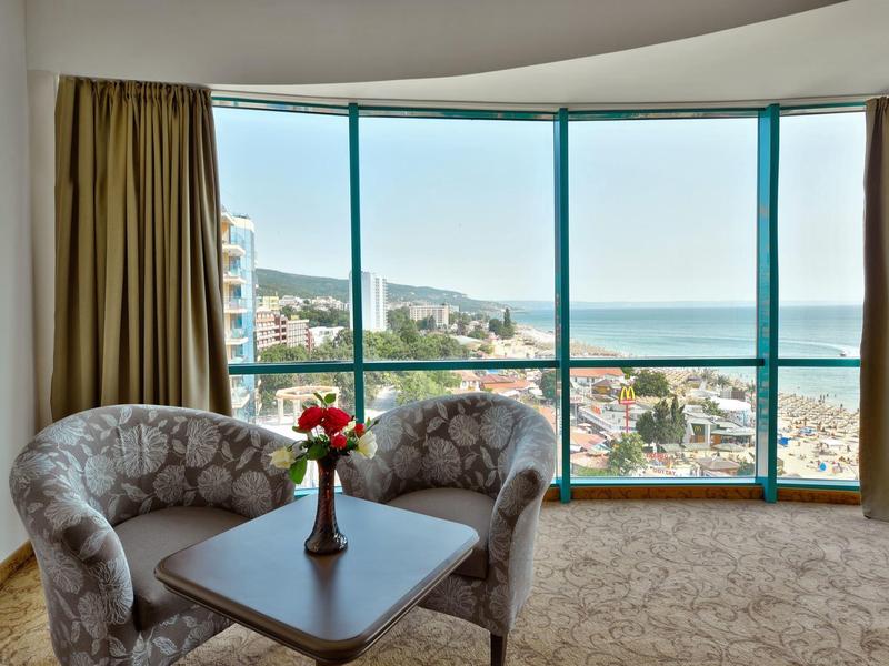 Pokój hotelowy z dwoma fotelami, stołem i dużymi oknami z widokiem na morze.