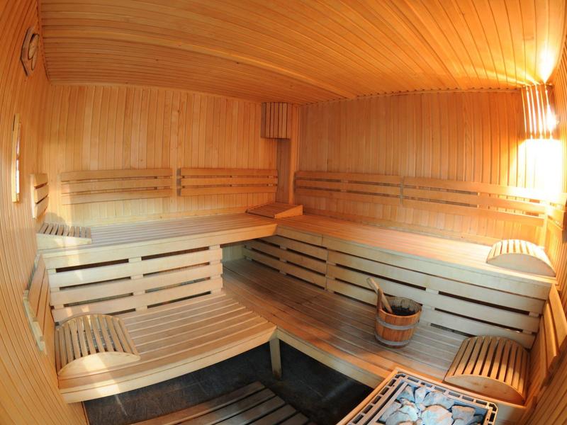 Jasno oświetlona sauna z drewnianymi ławkami, wiaderkiem i chochlą, lampami ściennymi i drewnianymi ścianami.