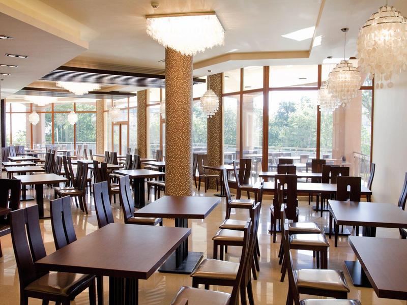 Leerer, heller Speisesaal mit vielen dunklen Holztischen und Stühlen, großen Fenstern und Kronleuchtern.