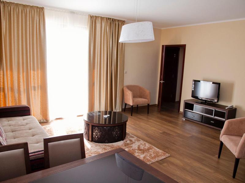 Hell eingerichtetes Wohnzimmer mit braunen Möbeln, Holzboden, TV und großen Fenstern mit Vorhängen.