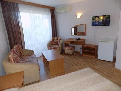 Piccola camera d'hotel con zona salotto, scrivania, mini frigorifero e finestra con tende.