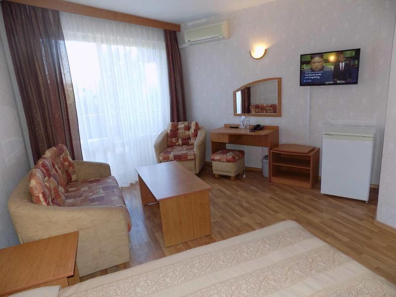 Piccola camera d'hotel con zona salotto, scrivania, mini frigorifero e finestra con tende.