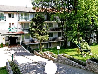 Widok zewnętrzny trzyklatkowego hotelu z wejściem, drzewami i zadbanym ogrodem.