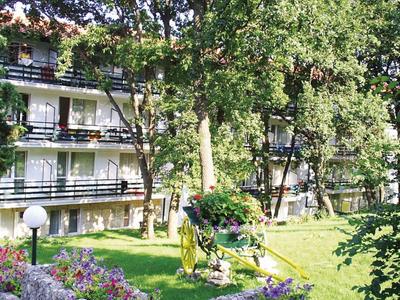 Vícepodlažní hotelová budova s balkony obklopená zelenými stromy a trávníky.