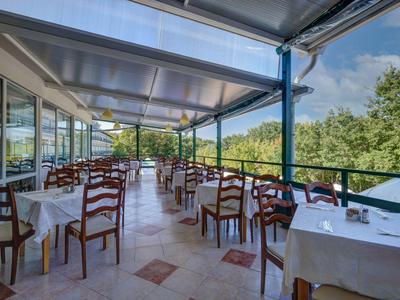 Zadaszony taras zewnętrzny z nakrytymi stołami i drewnianymi krzesłami z widokiem na zieleń.