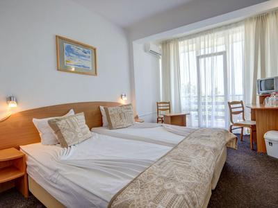 Světý hotelový pokoj se dvěma oddělenými postelemi, psacím stolem, židlemi a velkým oknem s bílou záclonou.