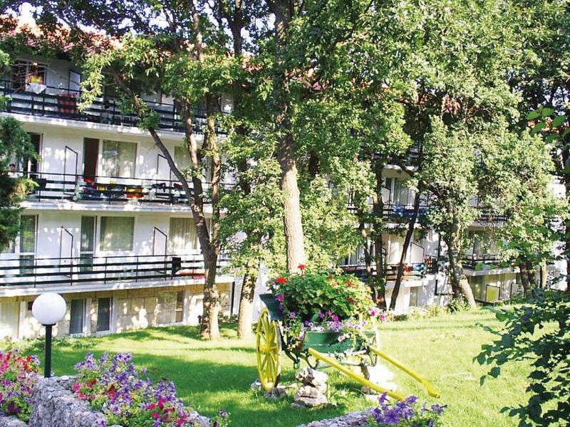 Vícepodlažní hotelová budova s balkony obklopená zelenými stromy a trávníky.