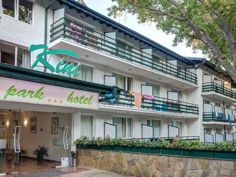 Vícepodlažní hotelová budova s balkony a zeleným označením, obklopená stromy.