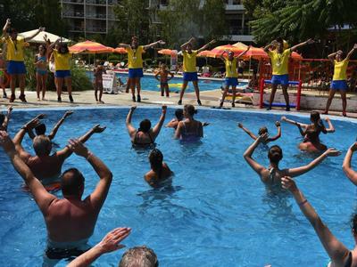 Lidé cvičí ve vodě v bazénu, instruktoři předvádějí cviky u okraje.