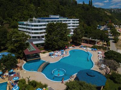 Hotel s velkým nepravidelně tvarovaným venkovním bazénem a lehátky, obklopen stromy.