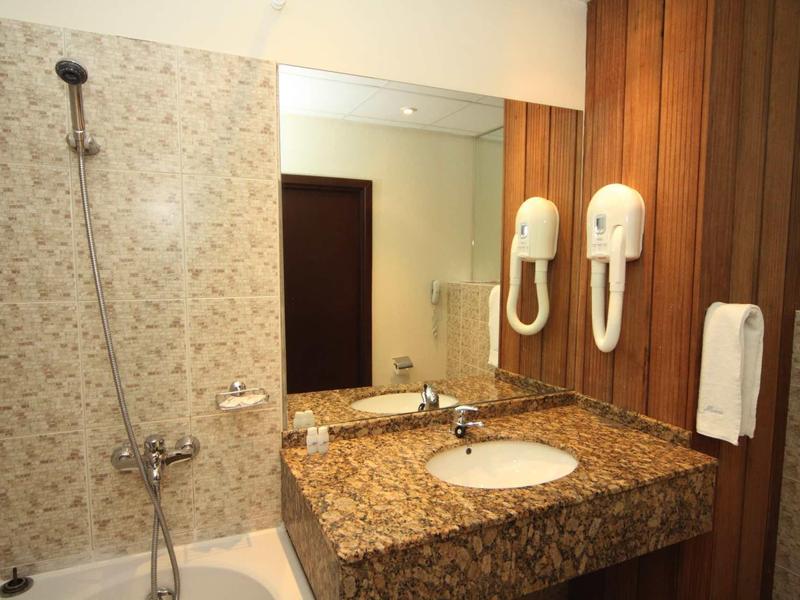 Hotelová koupelna se sprchou, velkým zrcadlem, granitovým umyvadlem a nástěnným fénem.