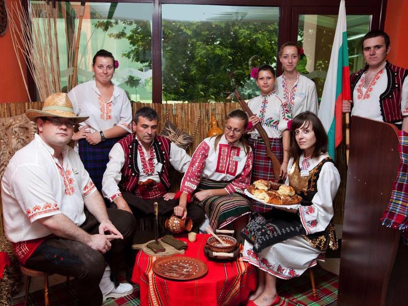 Skupina v tradičních krojích sedí v útulné místnosti s výhledem z okna.