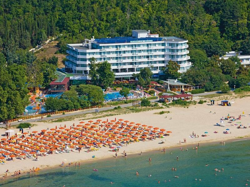 Velký hotel u zalesněné pláže s mnoha slunečníky a lidmi opalujícími se u moře.