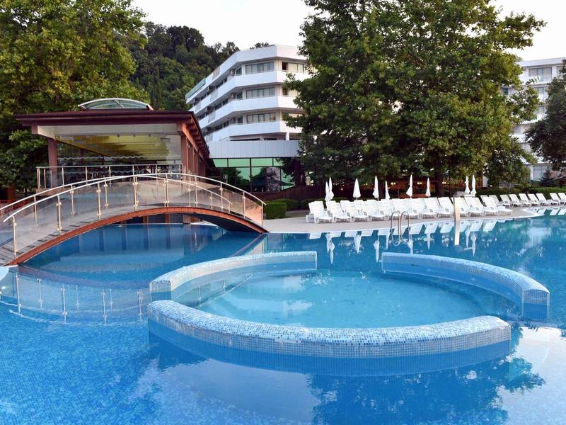 Bazén s mostkem, lehátky a budovou hotelu v pozadí.