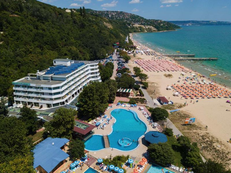 Hotel s modrým bazénem vedle dlouhé písečné pláže a zelených zalesněných útesů.