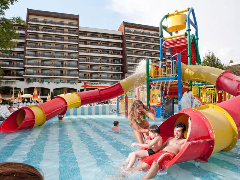 Děti sjíždějí barevné vodní skluzavky do bazénu před velkým hotelem.