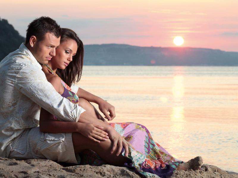 Pár sedí uvolněně na pláži a sleduje západ slunce nad mořem.