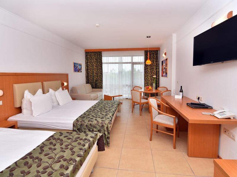 Světla hotelový pokoj se dvěma postelemi, TV na stěně a psacím stolem se židlemi.