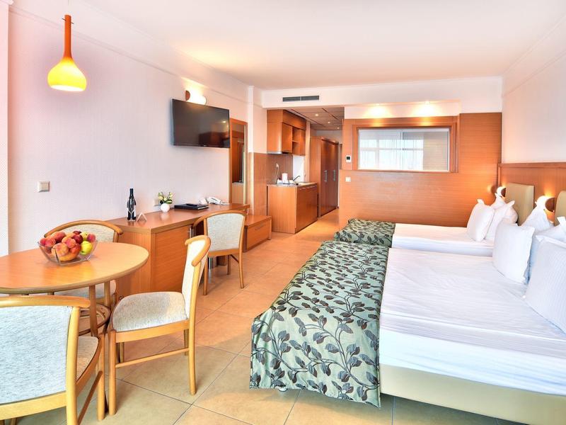 Světly hotelový pokoj s manželskou postelí, jídelním stolem se židlemi a moderním zařízením.