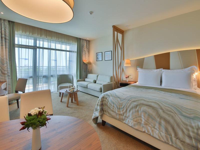 Světý hotelový pokoj s velkou postelí, pohovkou, psacím stolem a balkonem s výhledem ven