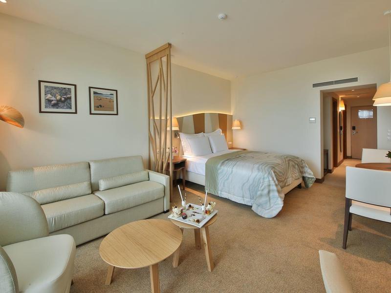 Moderně zařízený hotelový pokoj s manželskou postelí, pohovkou a jídelním stolem v neutrálních barvách.