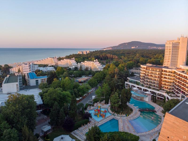 Pohled na hotel se tvarovaným bazénem, zelení a mořem při západu slunce.