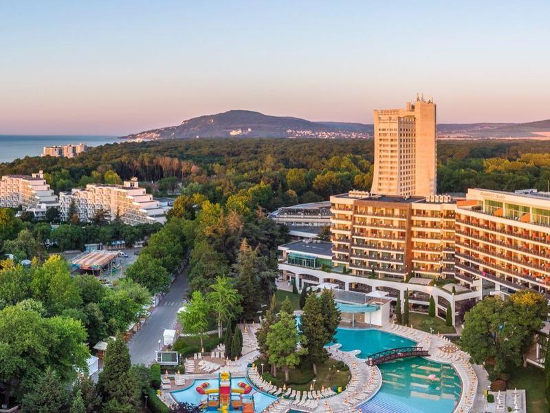 Panoramatický pohled na hotelový resort s bazény, obklopený zelenými stromy a horami v pozadí.