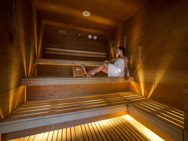 Osoba relaxuje v dřevěné sauně s víceúrovňovými lavičkami a osvětlením.