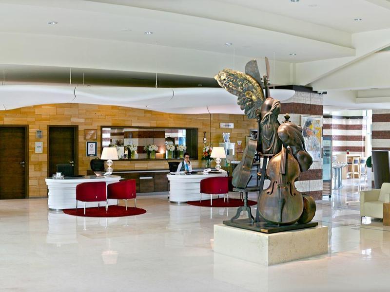 Světlá hotelová recepce s moderním nábytkem, rostlinami a velkou sochou uprostřed.